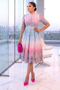A woman models a pink and blue lace midi dress with a high collar and short sleeves, holding a hot pink clutch and wearing hot pink heels in a modern glass-walled room.